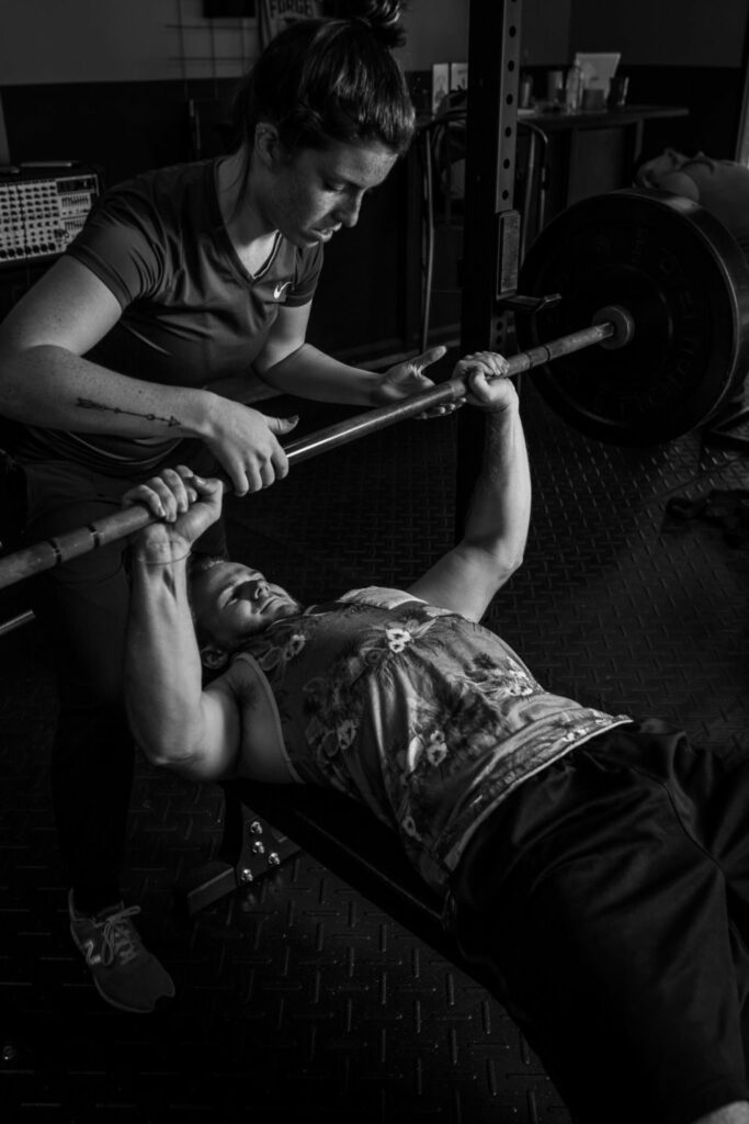 A personal trainer guiding a man as he lifts a barbell in a gym.