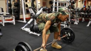 An old lady wearing camo shirt and bandana engaging in heavy lifting with a barbell in a gym.