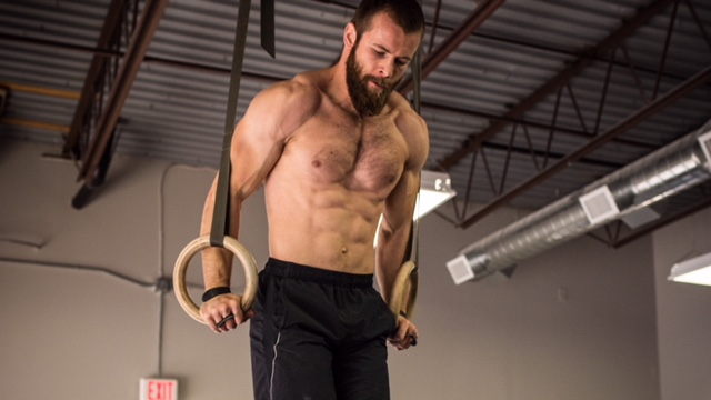 A bearded man is demonstrating his impressive core strength as he effortlessly performs rings exercises in a gym.