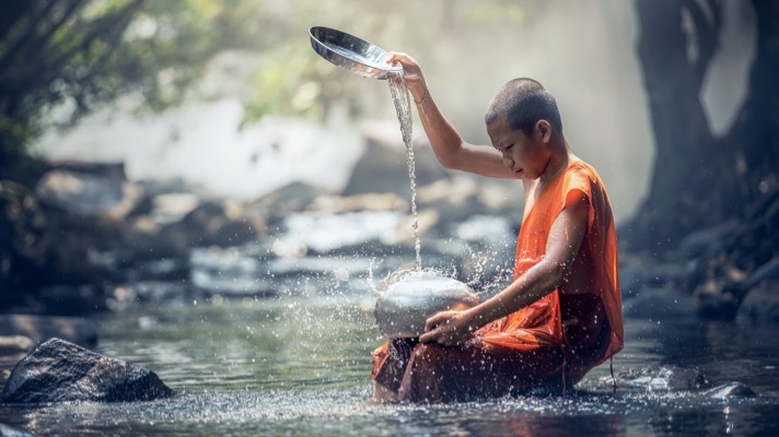 A monk in an orange robe is meditatively washing a pot in a serene river.